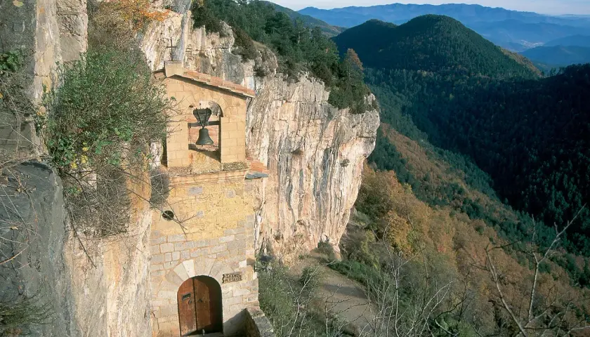 Ermita en la costa pubilla, aferrada a un acantilado rocoso con vistas a un valle boscoso. Una campana cuelga sobre la entrada.