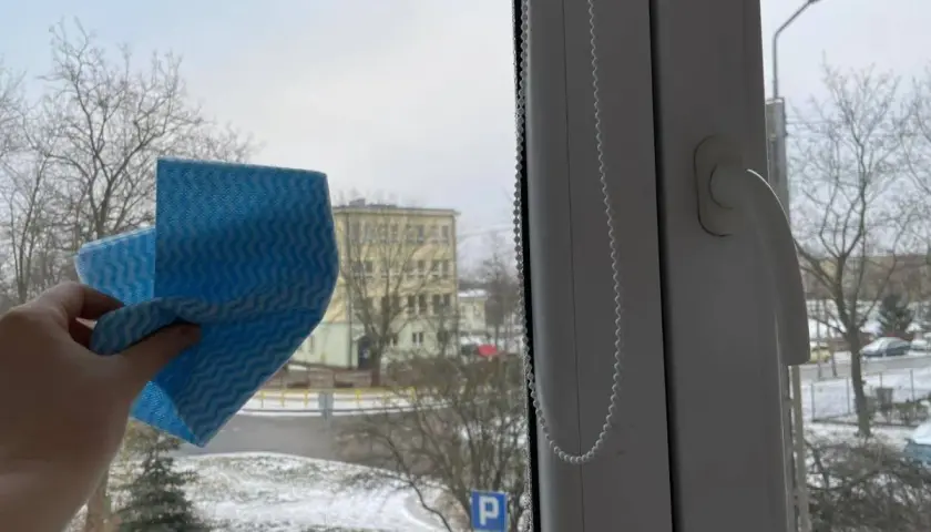 Ręka trzyma niebieską ściereczkę, gotową do mycia okna. Za szybą zimowy krajobraz z drzewami i budynkiem. Czy można myć okna na mrozie?