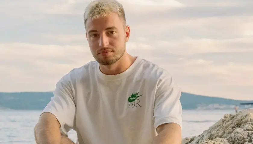 Edzio rap, z blond włosami i w białej koszulce, siedzi na plaży.