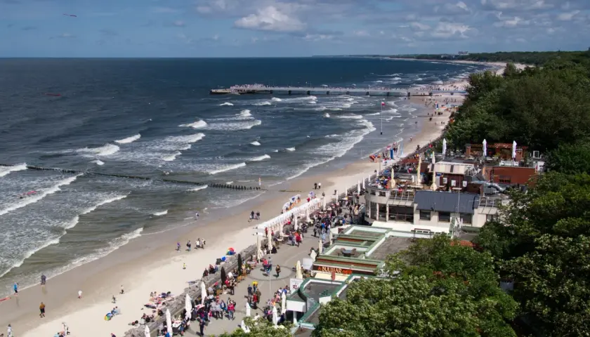 Tętniąca życiem promenada nad morzem, z plażą pełną ludzi, kawiarniami i molo w oddali.