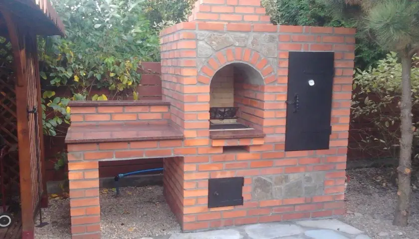 Solidny grill z cegły szamotowej, z kamiennym łukiem nad paleniskiem i metalowymi drzwiczkami. Idealny do ogrodu.
