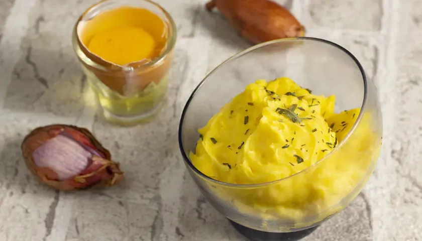 Una ciotola di salsa bernese cremosa, guarnita con erbe aromatiche, accanto a un tuorlo d'uovo e scalogni.