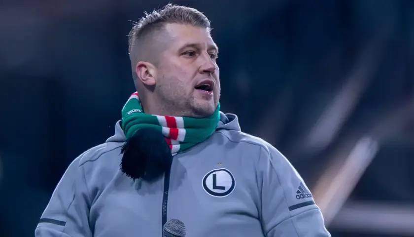 Staruch z Legii w szaliku klubowym, z mikrofonem przed ustami, gotowy do przemowy.
