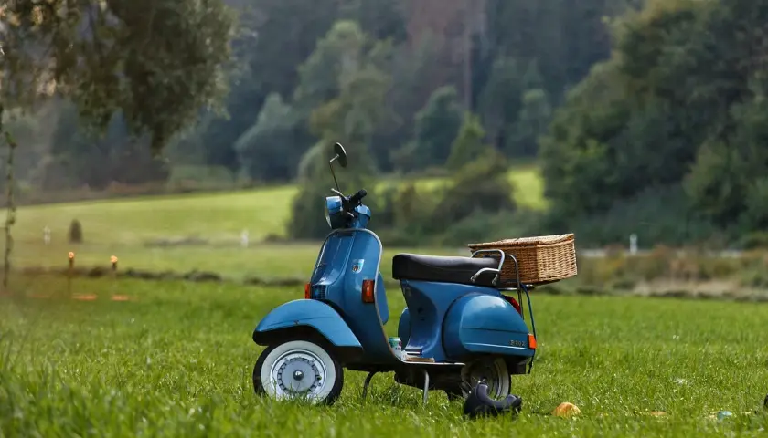 Niebieska Vespa z wiklinowym koszem na bagażniku, zaparkowana na trawie. Wygląda jakby czekała na przegląd skutera po terminie, gotowa na letnią przygodę.