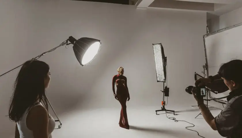 Modelka w czerwonej sukni pozuje na planie zdjęciowym. Fotograf z aparatem i asystentka przygotowują się do sesji.