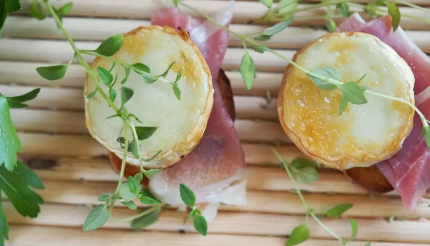 Dos bocaditos con crujiente de queso de cabra fundido y jamón serrano, adornados con hierbas frescas.