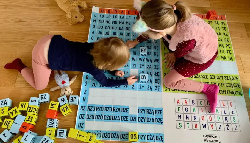 Dwie dziewczynki uczą się, jak nauczyć dziecko składać litery w wyraz, bawiąc się klockami z literami i planszą.