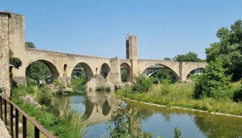 Puente medieval de Besalú, parte de una ruta histórica. Reflejo en el río y vegetación exuberante.