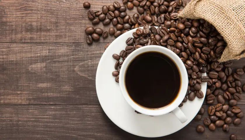 Aromatic coffee beans spill from a burlap sack onto a wooden table, next to a white cup filled with dark coffee. Poczuj kofeina działanie!