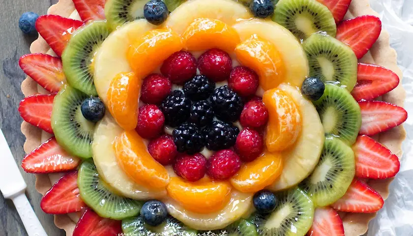 Torta alla frutta coloratissima, con fragole, kiwi, mandarini, lamponi e mirtilli disposti a raggiera su una base croccante.