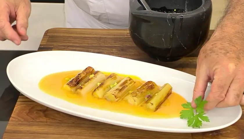 Puerros a la plancha, dorados y tiernos, servidos en una salsa anaranjada y decorados con perejil fresco.