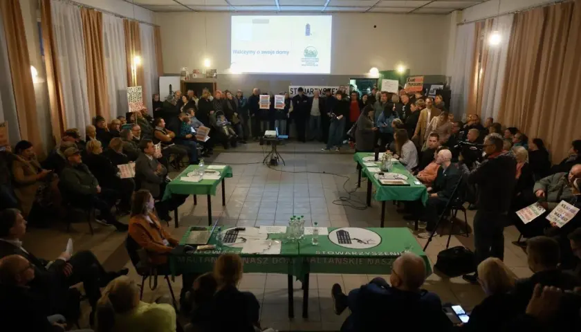 Ludzie siedzą i stoją na spotkaniu, na stole leżą dokumenty i laptop.