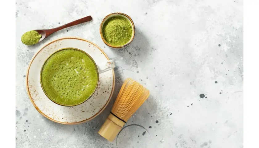 Jak pić herbatę matcha? Na stole leży spieniona zielona herbata w filiżance, obok bambusowa miotełka, łyżeczka z proszkiem i miseczka z matchą.