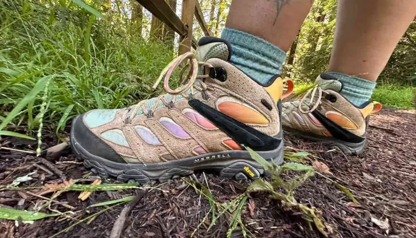 A person's feet wearing tan Merrell hiking boots with colorful accents and teal socks on a forest trail. These boots fit snugly, offering support for the hike.