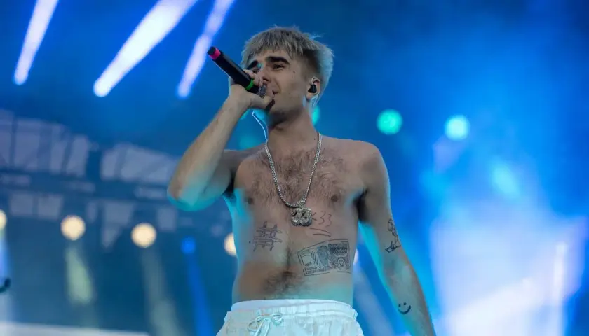 Artysta z tatuażami na klatce piersiowej, śpiewający do mikrofonu na koncercie. Jego styl przypomina okładki albumów.