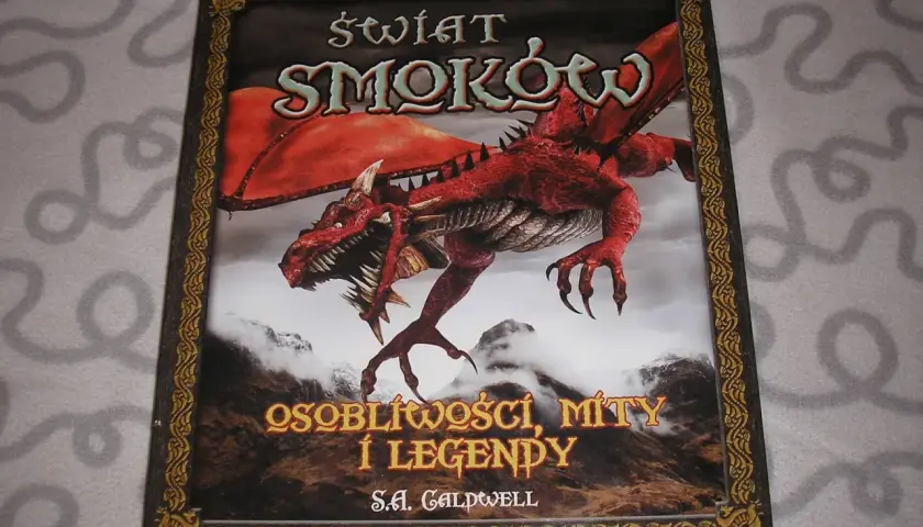 Okładka książki o smokach z czerwonym, groźnym stworzeniem na tle gór. Tytuł: "Świat Smoków: Osobliwości, Mity i Legendy".