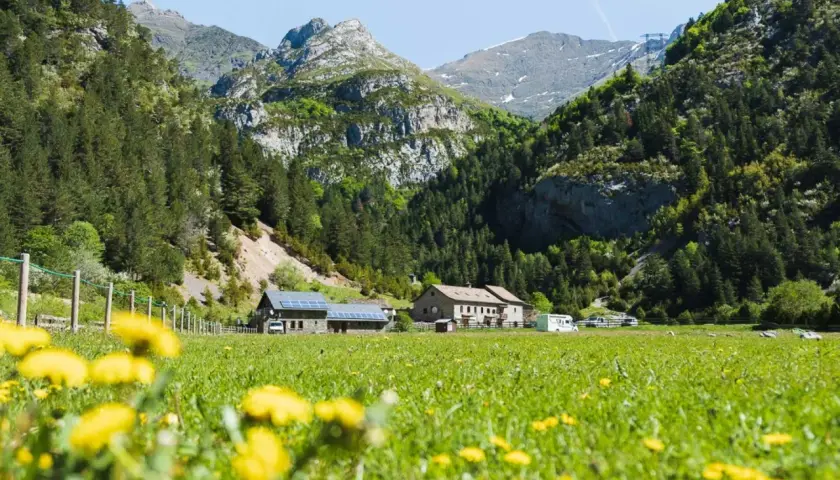 Prado florido con el Vignemale desde Bujaruelo al fondo. Casas rurales y montañas verdes bajo un cielo azul.