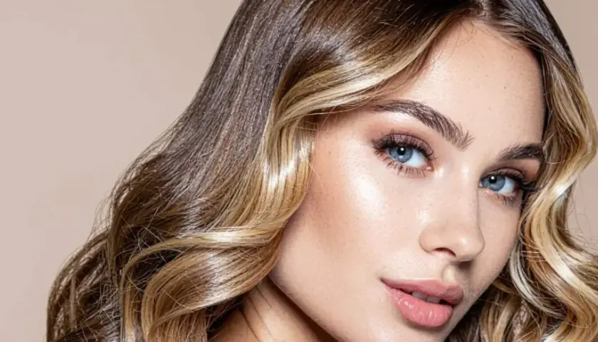 Mujer con cabello castaño y rubio, con un "money piece" que ilumina su rostro. Sus ojos azules y labios rosados resaltan su belleza.
