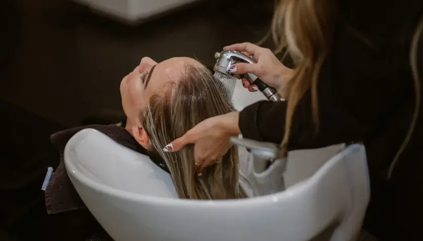 Una persona se lava el pelo con agua fría en un lavacabezas. El agua cae sobre su cabello rubio mientras una mano lo sujeta.