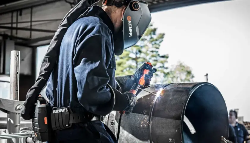 Spawacz wykonuje spawanie elektrodą w pionie na dużym metalowym elemencie. Widać iskry i łuk spawalniczy.