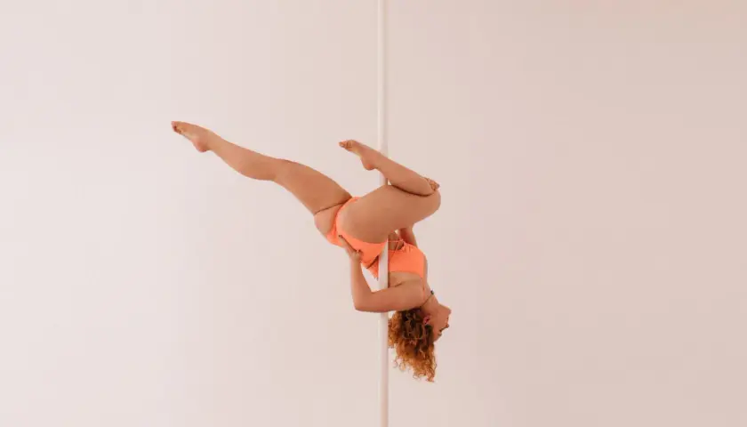 Kobieta w pomarańczowym stroju wykonuje skomplikowaną figurę na pionowym słupku, przypominającą flagę pole dance.