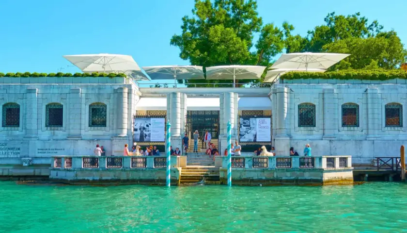 Le **Guggenheim Venise** se reflète dans l'eau turquoise. Des parasols blancs ombragent la terrasse où des visiteurs admirent l'architecture.