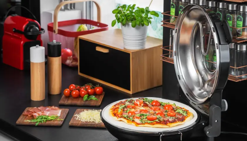Gotowa pizza na kamieniu w otwartym piecyku, obok składniki: pomidorki, rukola, szynka, ser.
