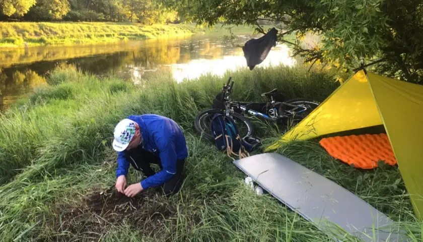 Biwak nad rzeką: rozłożony żółty namiot, materac, rower i mężczyzna przygotowujący ognisko. To idealny materiał na namiot i nocleg w plenerze.
