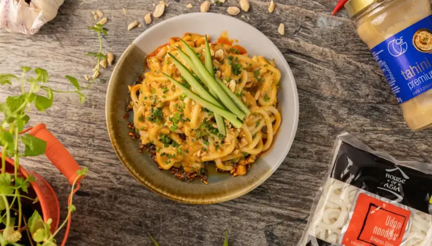 Pyszny makaron długi udon w kremowym sosie orzechowym, udekorowany ogórkiem i szczypiorkiem. Obok słoik tahini i paczka makaronu.