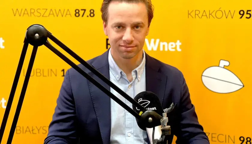 Krzysztof Bosak w garniturze przed mikrofonem na tle żółtej ściany z napisami.