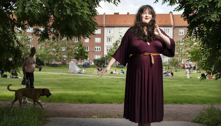 Maria Maunsbach i en vinröd klänning i en park med människor och en hund i bakgrunden.
