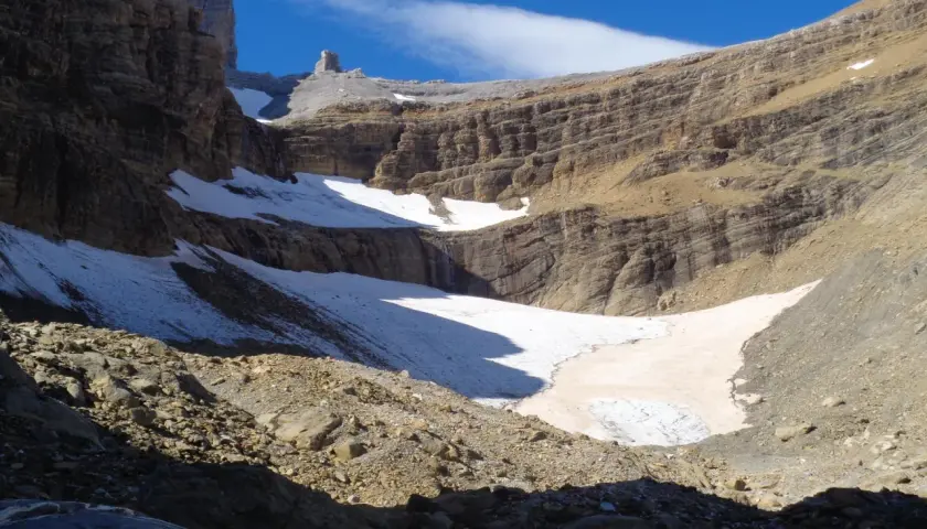 Un paisaje montañoso con nieve y rocas, donde el sol ilumina la **col de tentes** y el cielo azul.