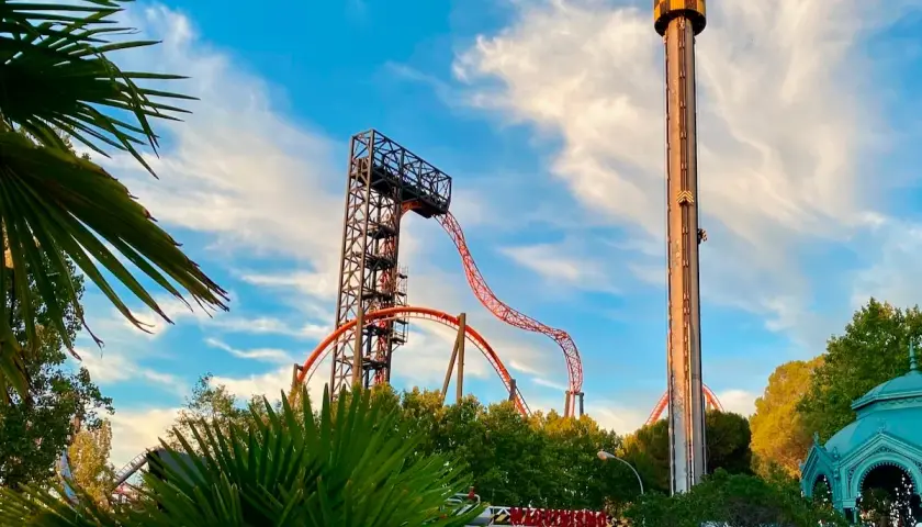 Torre de caída libre y montaña rusa anaranjada en un parque de atracciones con palmeras.
