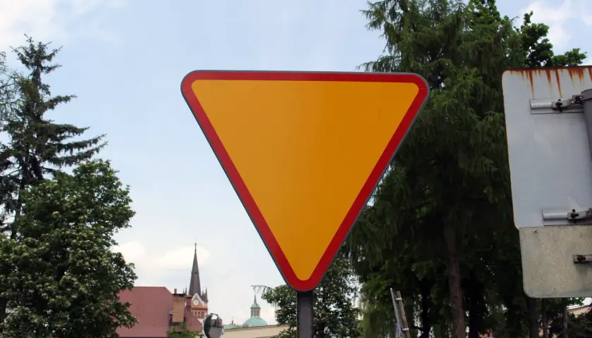Żółty, trójkątny znak ustąp pierwszeństwa z czerwoną obwódką, stojący na tle drzew i nieba.