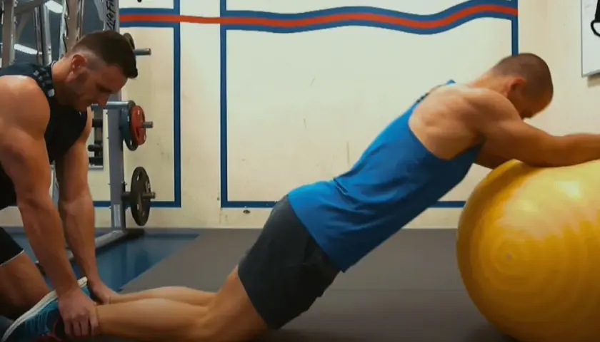 Trener pomaga wykonać ćwiczenie nordic curl, stabilizując nogi klienta opierającego się o dużą, żółtą piłkę gimnastyczną.