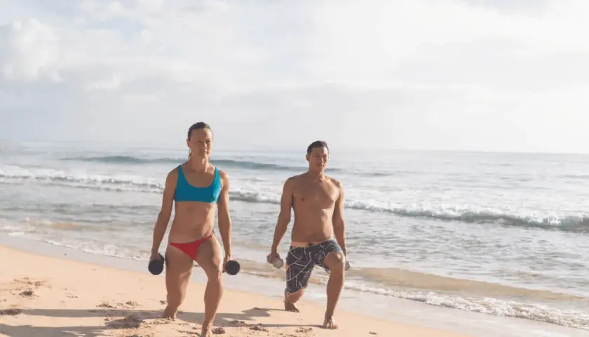 Kobieta i mężczyzna ćwiczą na plaży z hantlami. Ich ruchy to przykład świadomej kinezjologii, poprawiającej siłę i koordynację.
