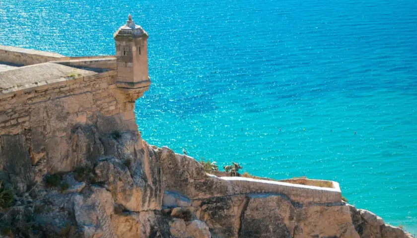 Torreón en el Castillo de Santa Bárbara, con el mar turquesa de fondo. Una vista icónica de la exposición alicantina.