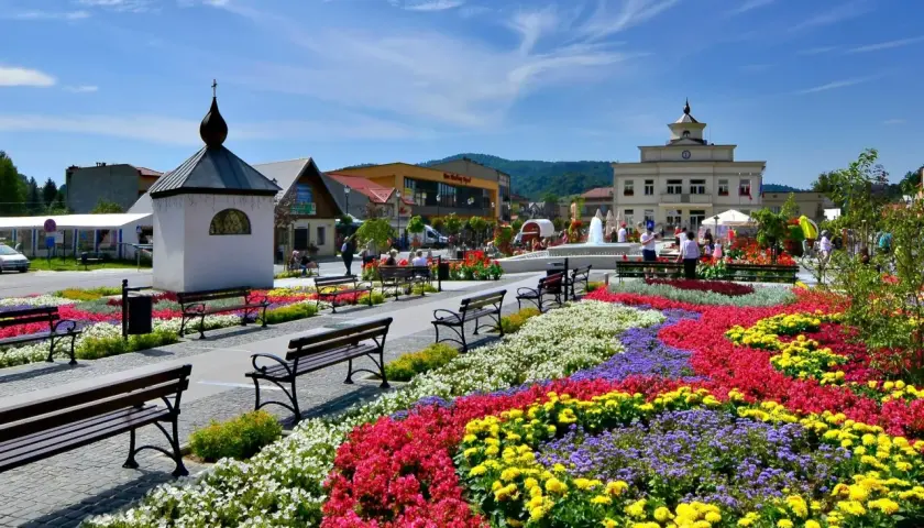 Kolorowe rabaty kwiatowe na rynku w Muszynie-Zdroju, z fontanną i budynkami w tle.