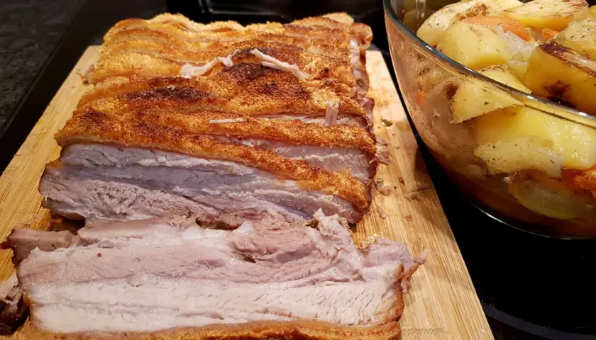 Ein butterzarter Schweinebauch mit knuspriger Kruste liegt auf einem Holzbrett, daneben eine Schüssel mit Bratkartoffeln und Gemüse.