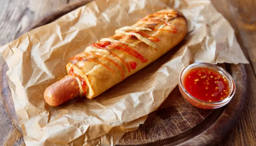Pyszny hot dog francuski w bułce, polany keczupem i posypany serem, z sosem na boku.