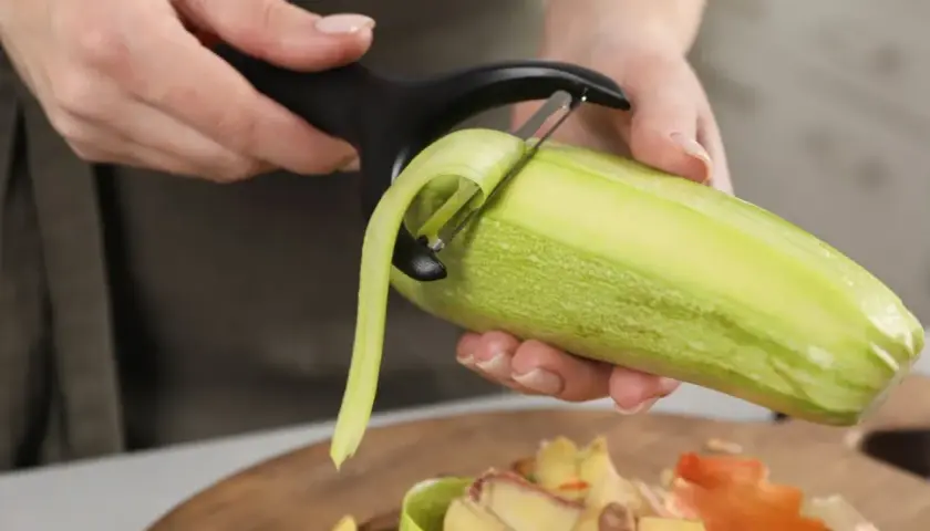 Une personne épluche une courgette. Les épluchures tombent sur une planche à découper, prêtes à être données aux poules.