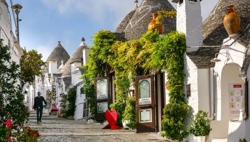 A person walks down a charming street lined with iconic trulli houses, lush greenery, and shops. This picturesque scene makes you wonder, which is the best town to stay in Puglia?