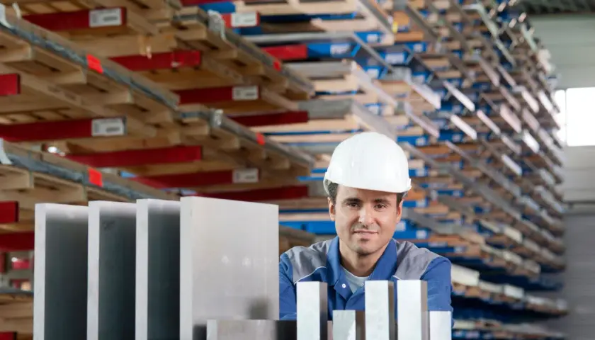 Pracownik w białym kasku stoi przed regałami pełnymi materiałów. Na pierwszym planie widać sztabki, które mogą być wykonane ze stopów aluminium.