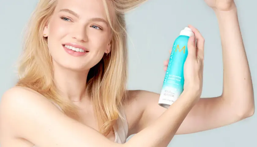 Mujer rubia sonriendo aplica champú seco para lavar pelo en seco, refrescando su melena.
