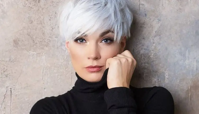 Kobieta z siwymi włosami w stylu pixie bob fryzura, ubrana w czarny golf, opiera dłoń o twarz.