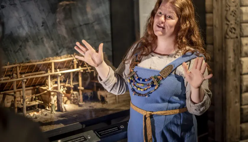 En kvinna i vikingakläder berättar vid en modell av en vikingagård på vikingamuseet.