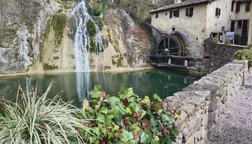 Antico mulino ad acqua accanto a cascate e grotte in Veneto, con un laghetto verde e vegetazione rigogliosa.