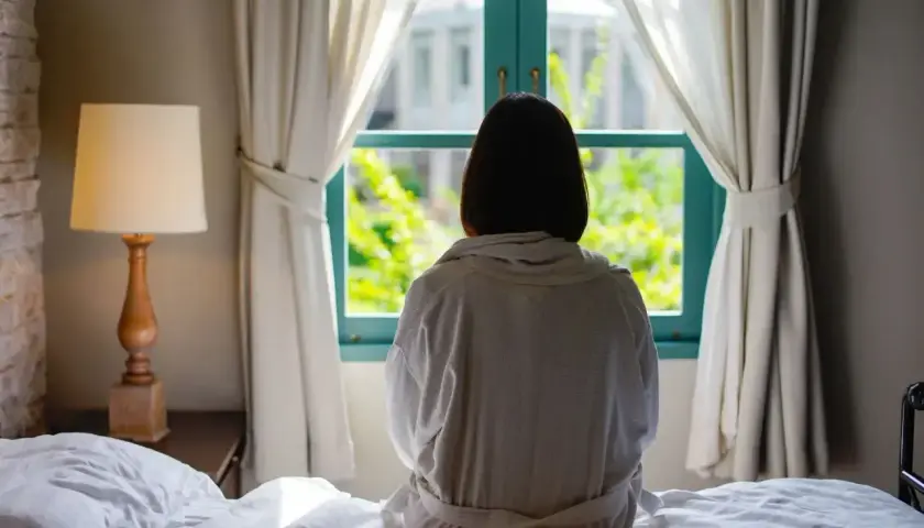 Osoba w szlafroku siedzi na łóżku, patrząc przez okno. Czasem czuje się jak w katatonii, bez ruchu, wpatrzona w zieleń.