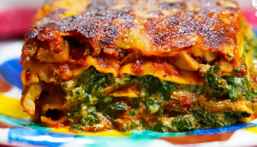 Porción de lasaña de verduras, con capas de pasta, espinacas y salsa roja, servida en un plato colorido.