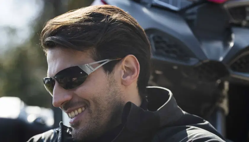 Uomo sorridente con occhiali da moto sportivi, con una moto sullo sfondo.